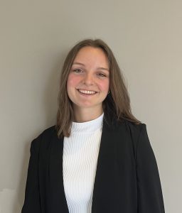 A woman with brown hair wearing a white turtle neck and dark blazer smiles into the camera.